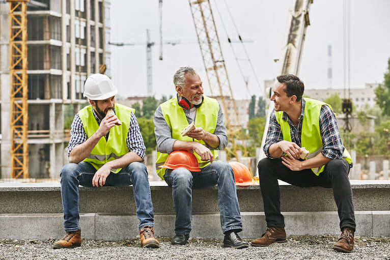 Group of construction workers eating sandwiches and talking while sitting on stone surface at construction site.